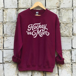 K&ouml;nnte beinhalten: Burgunderfarbener Sweatshirt mit wei&szlig;em Text, der "Hockey #66 Mom" in einer Retro-Schriftart lautet.