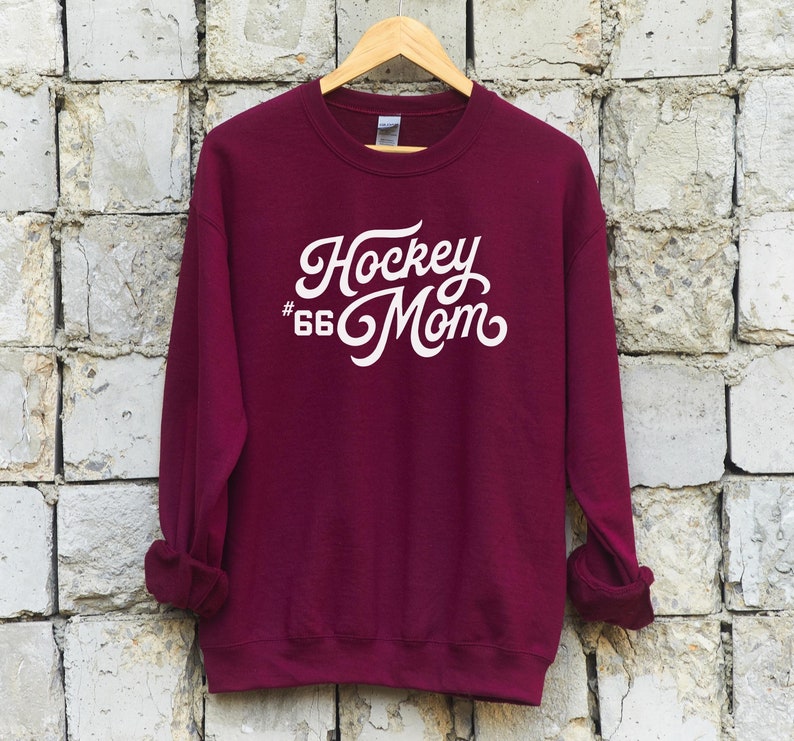 K&ouml;nnte beinhalten: Burgunderfarbener Sweatshirt mit wei&szlig;em Text, der "Hockey #66 Mom" in einer Retro-Schriftart lautet.
