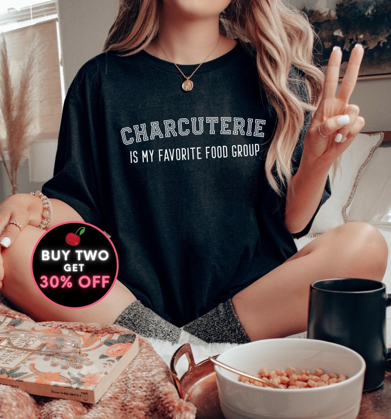 Peut inclure: Un t-shirt noir avec du texte blanc qui dit "Charcuterie is my favorite food group". Le t-shirt est port&eacute; par une personne aux cheveux blonds. Un autocollant rouge et blanc avec le texte "Buy Two Get 30% Off" se trouve dans le coin inf&eacute;rieur gauche de l'image.