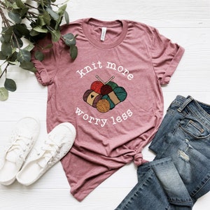 May include: A maroon t-shirt with the words "knit more worry less" in white, along with a graphic of yarn balls and knitting needles. The shirt is paired with blue jeans and white lace-up sneakers, creating a casual, craft-themed ensemble.