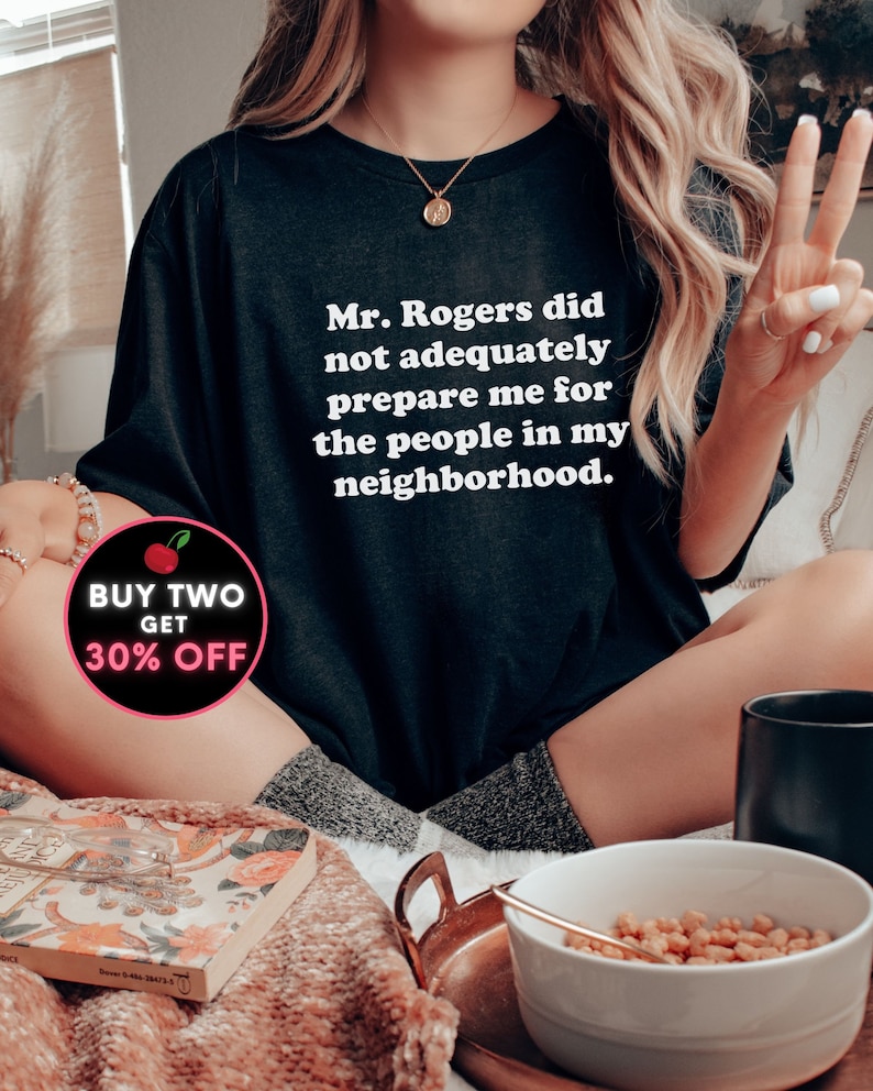 Puede incluir: Una camiseta negra con texto blanco que dice "Mr. Rogers did not adequately prepare me for the people in my neighborhood." Un gr&aacute;fico de cereza roja con el texto "BUY TWO GET 30% OFF" est&aacute; en la esquina inferior izquierda de la imagen.