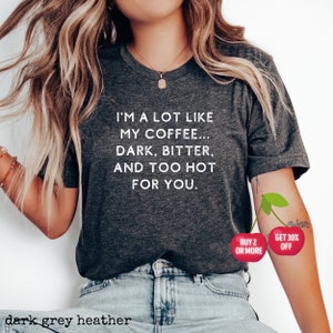 May include: Dark grey heather t-shirt with white text that reads "I'm a lot like my coffee... dark, bitter, and too hot for you."  The shirt is being modeled by a person wearing blue jeans. There are two red cherries on the person's arm with white text that reads "Buy 2 or more" and "Get 30% off".
