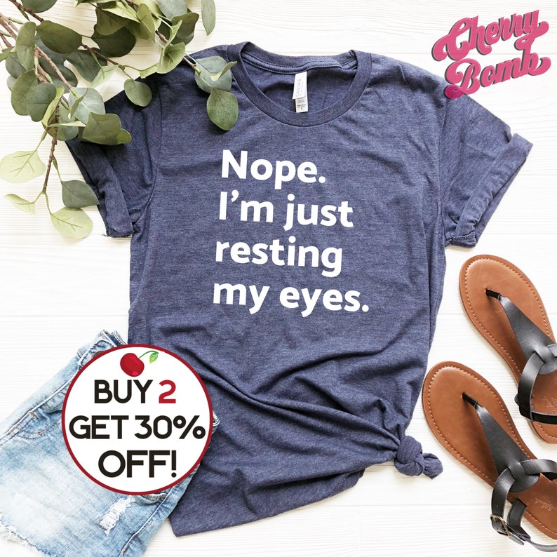 May include: A navy blue t-shirt with white text that reads "Nope. I'm just resting my eyes." The shirt is laying on a white surface with a pair of brown sandals and a pair of blue denim shorts. A red and white circle with a cherry graphic and the text "BUY 2 GET 30% OFF!" is in the lower left corner of the image.