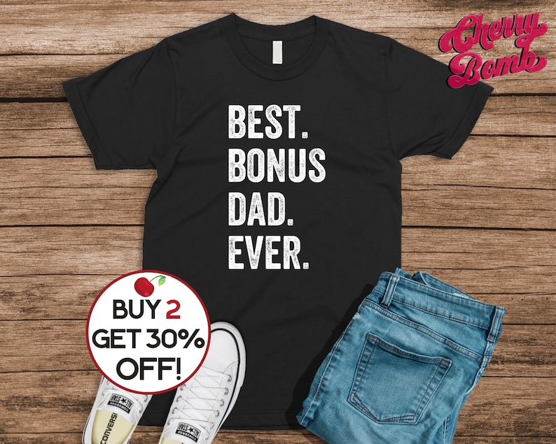 Puede incluir: Camiseta negra con el texto "BEST. BONUS DAD. EVER." en blanco. La camiseta se combina con vaqueros azules y zapatillas blancas. Un gr&aacute;fico circular dice "BUY 2 GET 30% OFF!"