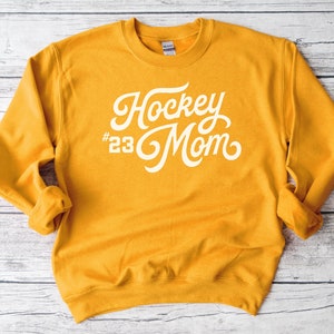 K&ouml;nnte beinhalten: Senfgelber Pullover mit wei&szlig;em Text, der in einer kursiven Schrift "Hockey #23 Mom" lautet.