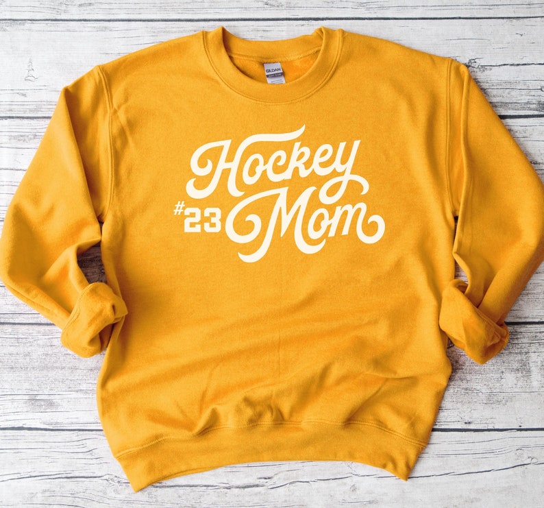 K&ouml;nnte beinhalten: Senfgelber Pullover mit wei&szlig;em Text, der in einer kursiven Schrift "Hockey #23 Mom" lautet.