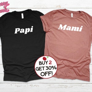 Peut inclure: Deux t-shirts avec les mots "Papi" et "Mami" imprim&eacute;s en blanc sur un t-shirt noir et un t-shirt rose. Les t-shirts sont sur un fond en bois blanc.  ACHETEZ 2, OBTENEZ 30% DE REMISE !