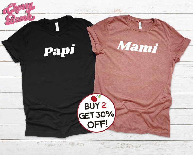 Peut inclure: Deux t-shirts avec les mots "Papi" et "Mami" imprim&eacute;s en blanc sur un t-shirt noir et un t-shirt rose. Les t-shirts sont sur un fond en bois blanc.  ACHETEZ 2, OBTENEZ 30% DE REMISE !