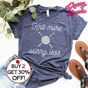 May include: A navy blue t-shirt with the text "Knit more worry less" in white. The text is accompanied by a graphic of a ball of yarn with two knitting needles crossed over it. The t-shirt has a distressed look and is made of a soft, comfortable material.
