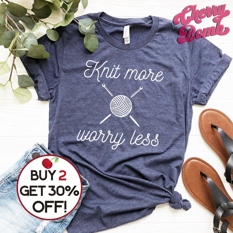 May include: A navy blue t-shirt with the text "Knit more worry less" in white. The text is accompanied by a graphic of a ball of yarn with two knitting needles crossed over it. The t-shirt has a distressed look and is made of a soft, comfortable material.