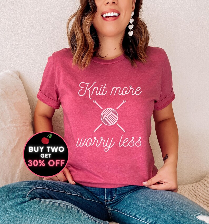 May include: A pink t-shirt with the text "Knit more worry less" in white. The text is accompanied by a graphic of a ball of yarn with knitting needles. A red circle with a white cherry and the text "BUY TWO GET 30% OFF" is in the lower left corner of the image.