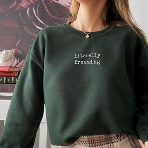 May include: Dark green sweatshirt with the text "literally freezing" in white, centered on the chest. The sweatshirt has a crew neck and long sleeves. The background features a floral design.