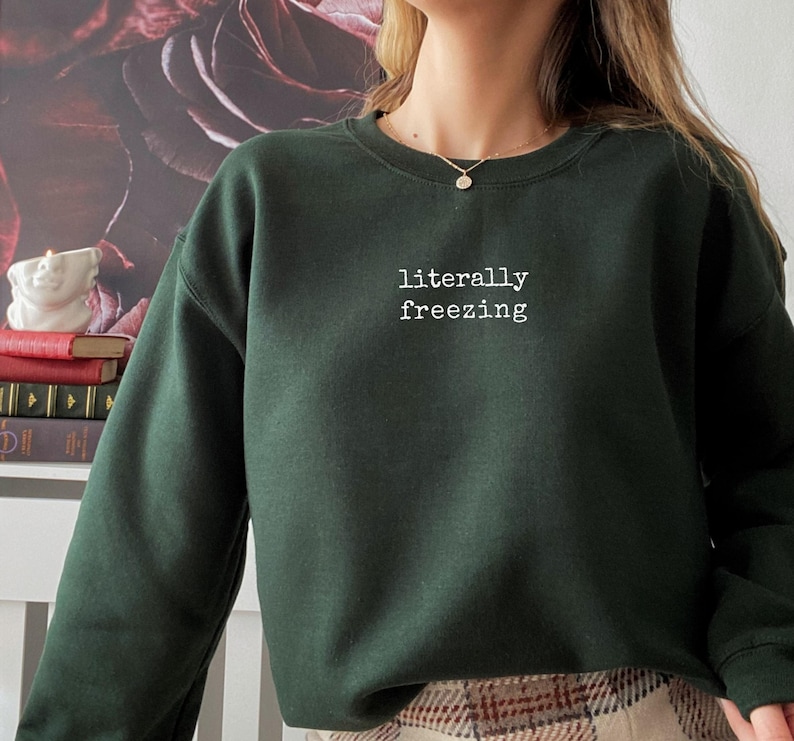May include: Dark green sweatshirt with the text "literally freezing" in white, centered on the chest. The sweatshirt has a crew neck and long sleeves. The background features a floral design.