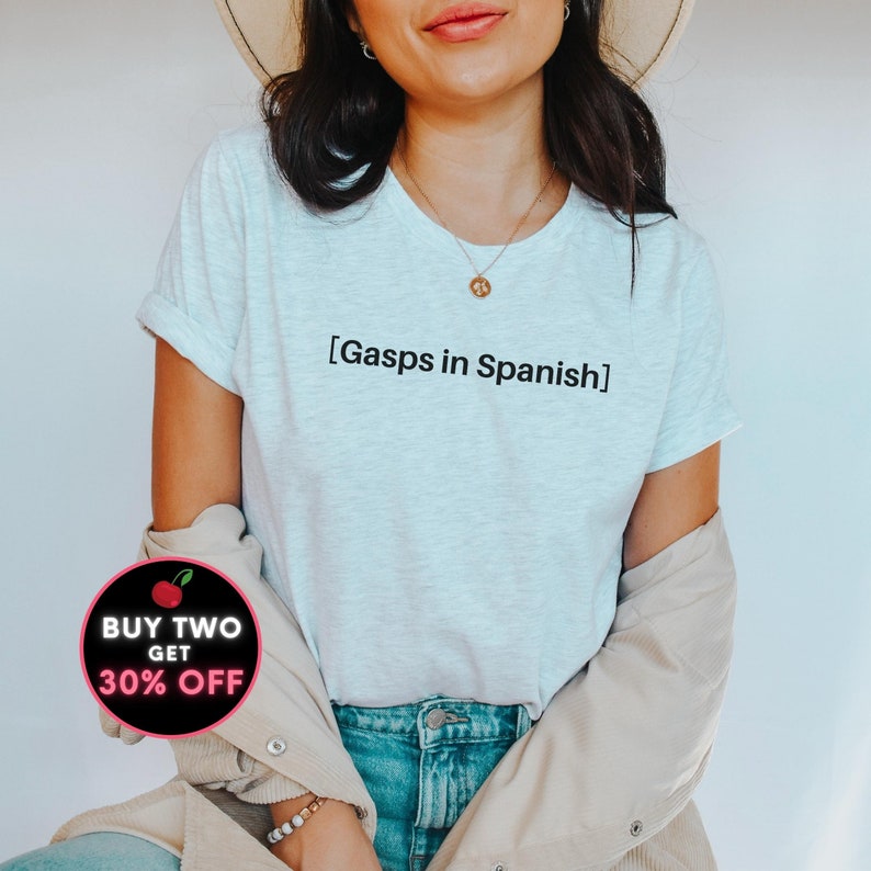 Peut inclure: Un t-shirt blanc avec le texte "[Gasps in Spanish]" imprim&eacute; sur le devant. Le t-shirt est port&eacute; par une personne aux cheveux longs bruns et un chapeau beige. La personne porte un jean bleu et une veste beige.