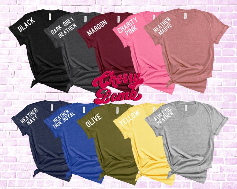 May include: A collection of ten t-shirts in various colors, including black, dark grey heather, maroon, charity pink, heather mauve, heather navy, heather true royal, olive, yellow, and athletic heather. Each shirt has a different color and a text label on the sleeve.