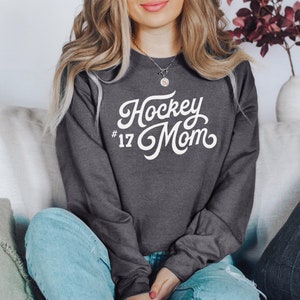 K&ouml;nnte beinhalten: Ein dunkelgrauer Pullover mit wei&szlig;em Text, der "Hockey Mom #17" in einer Retro-Schriftart lautet.