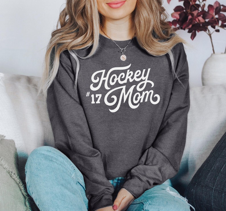 K&ouml;nnte beinhalten: Ein dunkelgrauer Pullover mit wei&szlig;em Text, der "Hockey Mom #17" in einer Retro-Schriftart lautet.