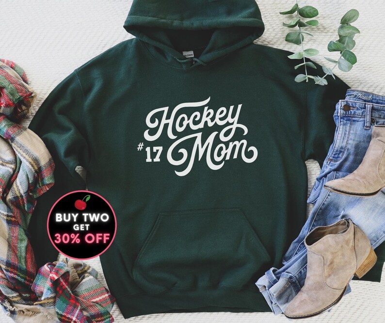 Puede incluir: Una sudadera con capucha verde oscuro con las palabras "Hockey Mom #17" en blanco. La sudadera tiene capucha y bolsillo delantero. La imagen tambi&eacute;n incluye unos vaqueros azules, una bufanda y un par de botas.