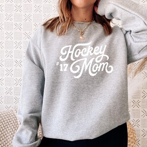 K&ouml;nnte beinhalten: Grauer Sweatshirt mit dem Text "Hockey Mom #17" in wei&szlig;er Schrift.
