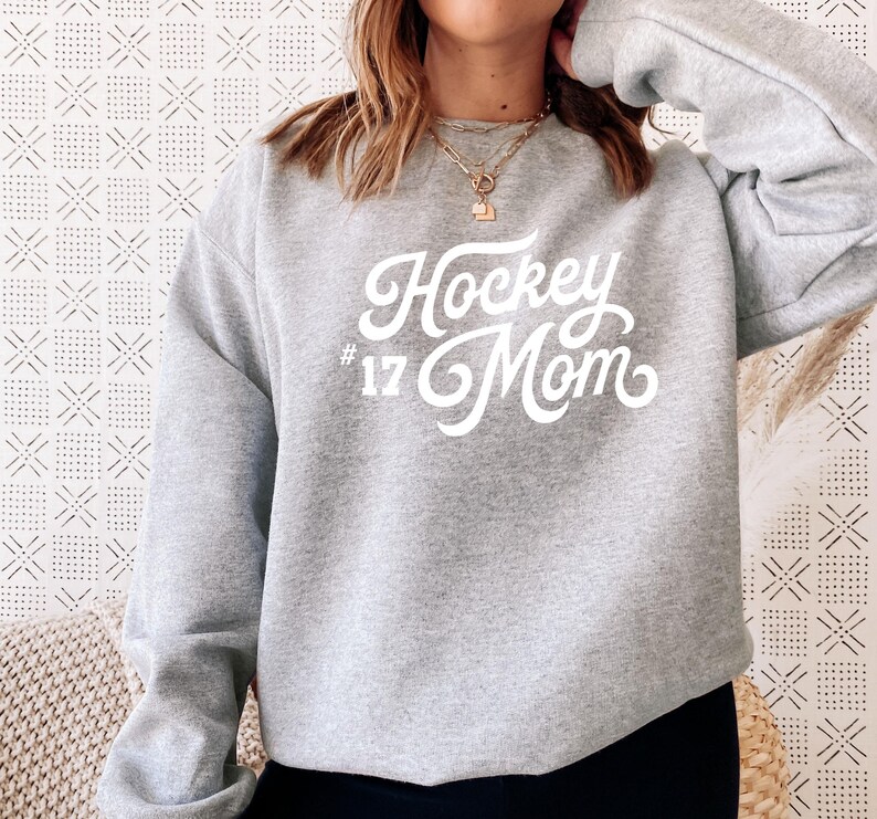 K&ouml;nnte beinhalten: Grauer Sweatshirt mit dem Text "Hockey Mom #17" in wei&szlig;er Schrift.