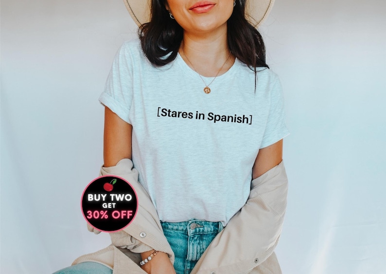 以下が含まれることがあります： 前面に「[Stares in Spanish]」という文字がプリントされた白いTシャツ。長い茶色の髪とベージュのジャケットを着た人が着用しています。画像の左下には、「BUY TWO GET 30% OFF」という文字が書かれたピンクと黒の円があります。