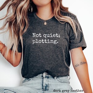 May include: Dark grey heather t-shirt with the text "Not quiet; plotting." in white. The shirt has a crew neck and short sleeves. The bottom of the shirt is visible, along with the top of a pair of blue jeans.