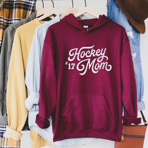 Puede incluir: Una sudadera con capucha de color granate con el texto "Hockey Mom #17" en escritura blanca. La sudadera tiene un bolsillo delantero y una capucha. Otras camisas y un par de vaqueros cuelgan en un estante en el fondo.