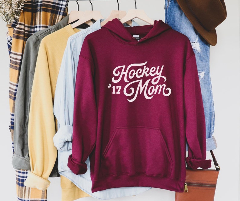 Puede incluir: Una sudadera con capucha de color granate con el texto "Hockey Mom #17" en escritura blanca. La sudadera tiene un bolsillo delantero y una capucha. Otras camisas y un par de vaqueros cuelgan en un estante en el fondo.