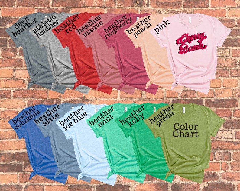 May include: A collection of various colored t-shirts, including heather gray, red, pink, and green. One shirt features the text "Cherry Bomb" in a retro font. Another shirt displays a "Color Chart" label. The shirts are displayed against a brick wall.
