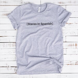 以下が含まれることがあります： グレーのTシャツに「[Stares in Spanish]」という文字がプリントされています。