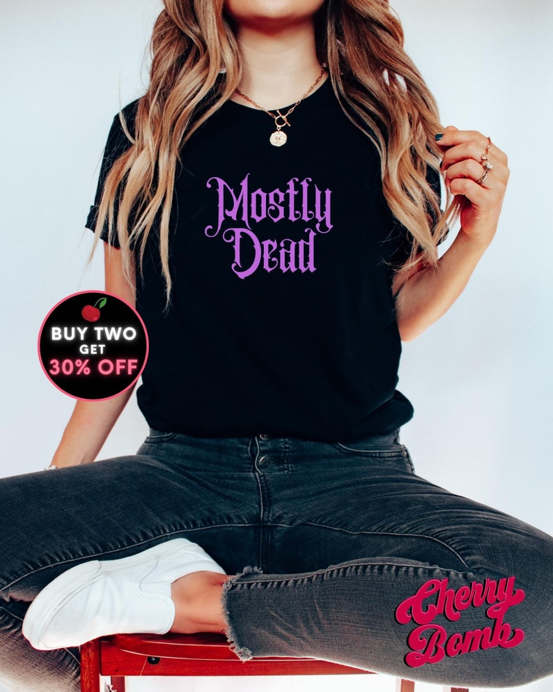 May include: A black t-shirt with the text "Mostly Dead" in purple script. The shirt is being worn by a person with long blonde hair and is sitting on a red chair. The text "BUY TWO GET 30% OFF" is in a red circle with a cherry graphic.