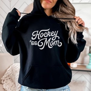 Puede incluir: Sudadera con capucha negra con bolsillo delantero y capucha con cord&oacute;n. El texto blanco en la parte delantera dice "Hockey Mom" con el n&uacute;mero "66" debajo. Es una prenda informal.