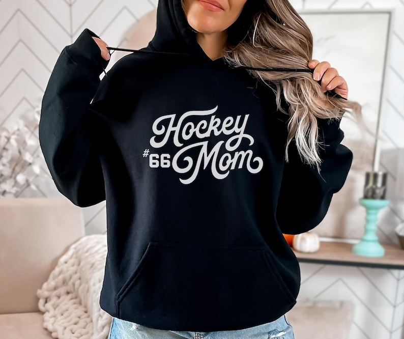 Puede incluir: Sudadera con capucha negra con bolsillo delantero y capucha con cord&oacute;n. El texto blanco en la parte delantera dice "Hockey Mom" con el n&uacute;mero "66" debajo. Es una prenda informal.