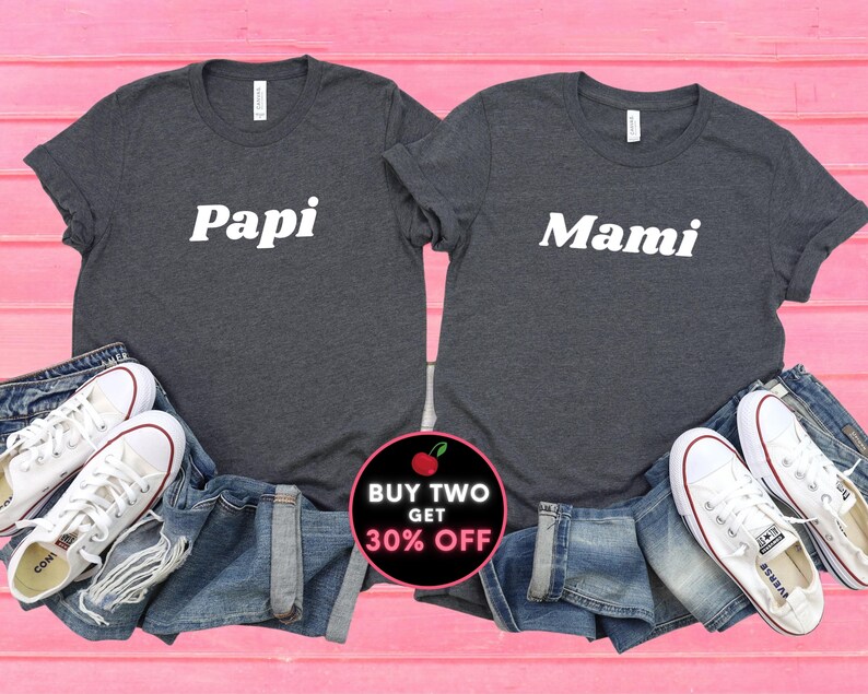 Peut inclure: Deux t-shirts gris fonc&eacute; avec du texte blanc. Le t-shirt de gauche dit "Papi" et celui de droite dit "Mami". Les t-shirts sont dispos&eacute;s sur une surface rose avec un jean bleu et des baskets blanches. Un cercle rouge et noir avec le texte "ACHETEZ DEUX, OBTENEZ 30% DE REMISE" se trouve dans le coin inf&eacute;rieur droit.