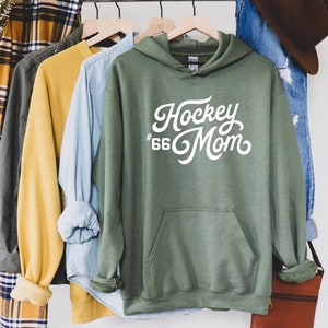 Puede incluir: Una sudadera con capucha verde con el texto blanco "Hockey Mom" y el n&uacute;mero "66". La sudadera tiene un bolsillo delantero y est&aacute; colgada en una percha de madera con otras camisas. Las mangas est&aacute;n enrolladas.