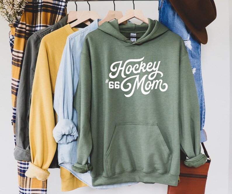 Puede incluir: Una sudadera con capucha verde con el texto blanco "Hockey Mom" y el n&uacute;mero "66". La sudadera tiene un bolsillo delantero y est&aacute; colgada en una percha de madera con otras camisas. Las mangas est&aacute;n enrolladas.
