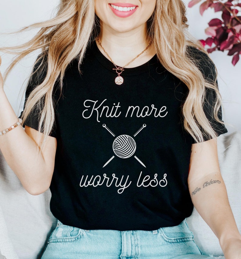 May include: A black t-shirt with the text "Knit more worry less" in white. The text is accompanied by a graphic of a ball of yarn with knitting needles.