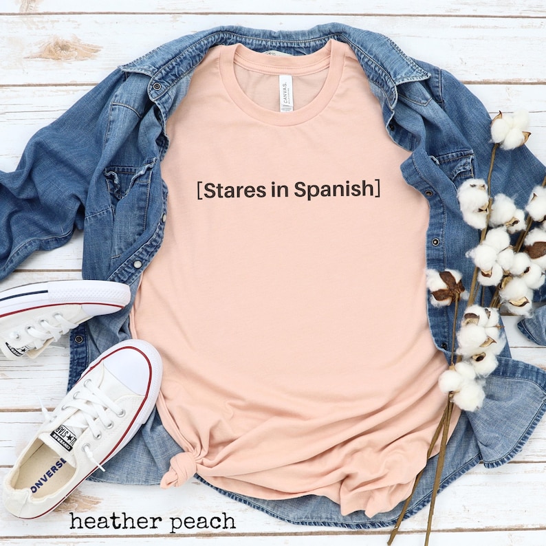 以下が含まれることがあります： 前面に「[Stares in Spanish]」という文字がプリントされた、ピーチ色のTシャツです。