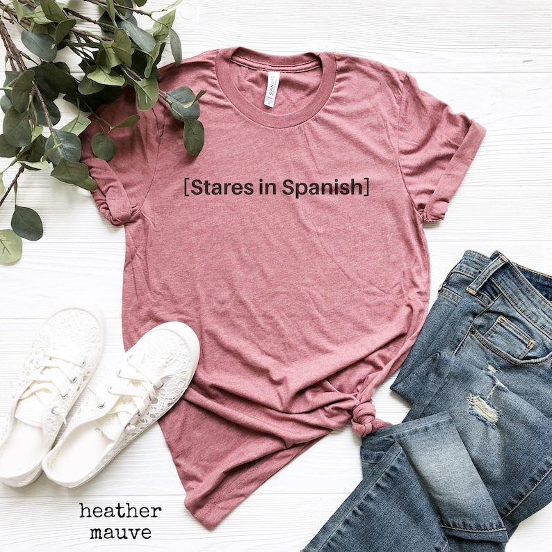以下が含まれることがあります： 正面に「[Stares in Spanish]」という文字がプリントされたヘザーモーヴのTシャツ。白い表面に白いレースアップシューズと青いジーンズと一緒に平らに置かれています。
