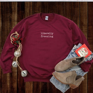 May include: A maroon sweatshirt with the text "literally freezing" in white. The sweatshirt is paired with blue jeans, tan ankle boots, and a holiday card. Decorative bells and greenery are also present, suggesting a winter or holiday theme.