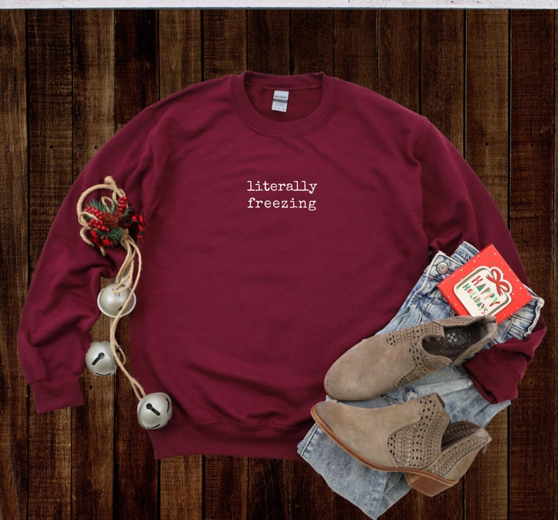 May include: A maroon sweatshirt with the text "literally freezing" in white. The sweatshirt is paired with blue jeans, tan ankle boots, and a holiday card. Decorative bells and greenery are also present, suggesting a winter or holiday theme.