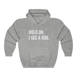 Pode incluir: Camisola de moletom cinza com capuz com o texto "HOLD ON, I SEE A DOG." impresso em branco.