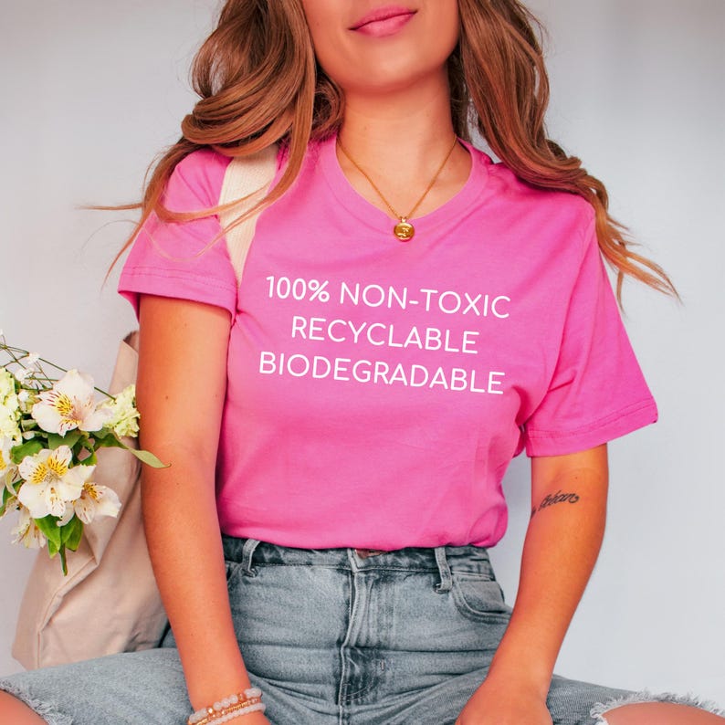 May include: A bright pink V-neck t-shirt with the words "100% NON-TOXIC RECYCLABLE BIODEGRADABLE" printed in white. The shirt is paired with blue denim jeans.