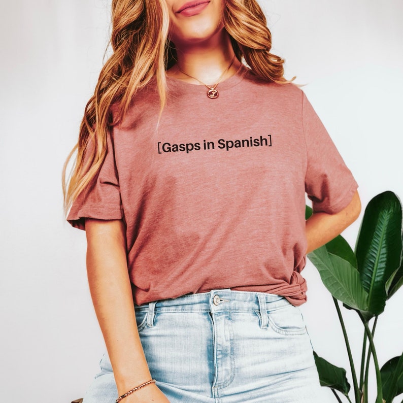 Peut inclure: Une femme portant un t-shirt mauve avec le texte "[Gasps in Spanish]" imprim&eacute; dessus. Elle porte un short en jean bleu clair et a de longs cheveux blonds.