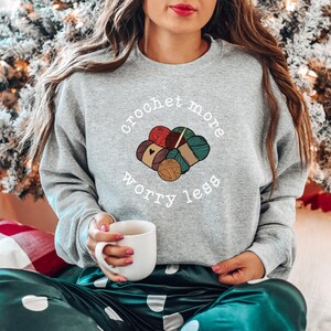 May include: A gray sweatshirt with the text "crochet more worry less" in white. The text is surrounded by a graphic of colorful yarn balls and knitting needles.