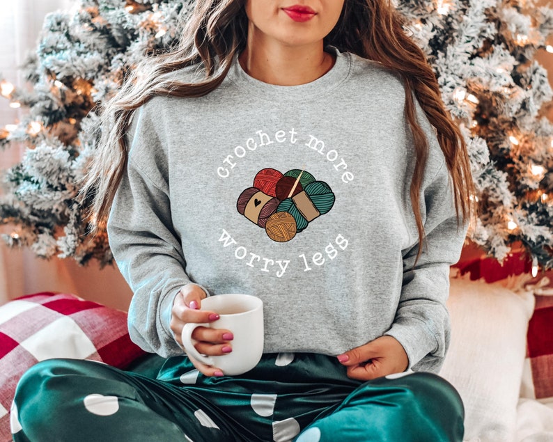 May include: A gray sweatshirt with the text "crochet more worry less" in white. The text is surrounded by a graphic of colorful yarn balls and knitting needles.