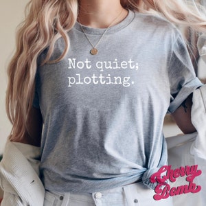 May include: A heather gray t-shirt with the text "Not quiet; plotting." in white. The shirt is knotted at the waist. The Cherry Bomb logo is visible in the bottom right corner.