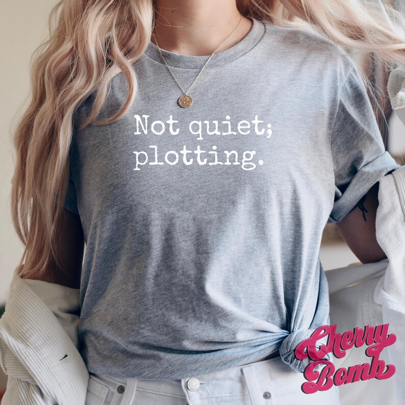 May include: A heather gray t-shirt with the text "Not quiet; plotting." in white. The shirt is knotted at the waist. The Cherry Bomb logo is visible in the bottom right corner.