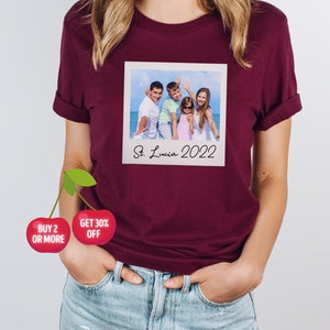 May include: Burgundy t-shirt featuring a printed image of a family on a beach, resembling a Polaroid photo. The photo is labeled "St. Lucia 2022". The shirt also has a cherry graphic with the text "BUY 2 OR MORE" and "GET 30% OFF".