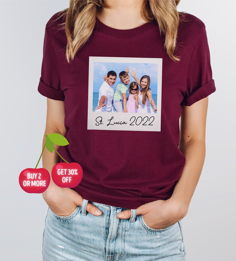 May include: Burgundy t-shirt featuring a printed image of a family on a beach, resembling a Polaroid photo. The photo is labeled "St. Lucia 2022". The shirt also has a cherry graphic with the text "BUY 2 OR MORE" and "GET 30% OFF".